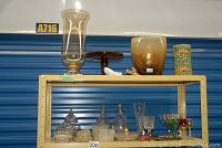 Glassware including tall hurricane glass, wooden pot stand, a large amber glass vase, and a green patterned vase with two sets of liqueur glasses including colored stem glasses on a shelf.