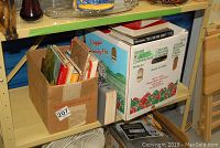 Boxes with assorted books including cookbooks, art reference, and non-fiction in cardboard boxes on a shelving unit.