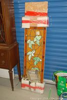 Full view of large stained glass panel wrapped with protective foam on top and bottom, leaning on MDF shelf with other items around it
