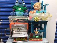 Blue metal Jack n Jill Kiddie Chair with doll on it, stuffed Stitch toy in green sweater, plastic craft supplies, and cast metal Trick Dog clown bank in front.