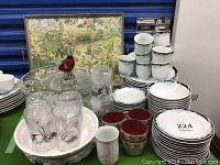 Overview of dishes, glasses, red Murano glass duck on plate, stacked china plates, and ceramic bowls in front with a colorful framed tray at the back.