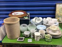 Photo showing china dishes including cups, saucers, bowls, plates and assorted mugs on a green table, stoneware crock, framed collector plate
