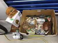 Box containing various small household and decorative items including figurines, costume jewelry in cedar box, coasters, and miscellaneous items.