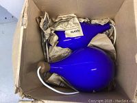 Two cobalt blue glass lamps packed in a cardboard box with newspaper wrapping, showing wood tops and hanging wires.