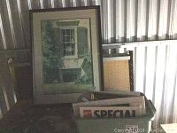 Framed print leaning against storage wall with box of advertising prints in front