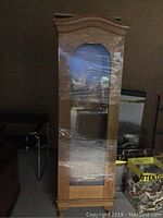 Full view of the oak cabinet showing the front glass door and curved top detail, wrapped in protective plastic.