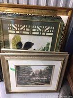 Two framed prints stacked against a wall, showing the landscape print with a house surrounded by trees in the front and the panda print partially visible behind.