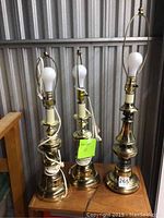 Three lamps standing on wooden surface, one taller brass base lamp and two shorter matching metal base lamps, no shades, with cords and bulbs attached.