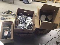 Four boxes with electronic items including cables, adapters, power strips, speakers, and media storage.