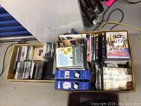 Photo of two cardboard boxes filled with DVDs, CDs, and two boxed phones. DVDs and CDs are stacked and arranged loosely. Two blue and white phone boxes placed inside the box.