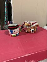 Two ceramic Christmas mugs on red surface, snowman and bear designs