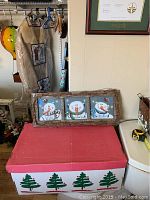 Overall view of the snowman wall decor placed on a red box; the frame and three snowman panels are visible