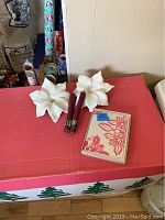 Wide shot showing two white poinsettia candle holders, two red taper candles, and a Hawaiian address book on a red surface, with wrapping paper rolls in the background.