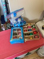 Wide view showing two boxes of vintage glass Christmas ball ornaments and a plastic bag of blue and silver ornaments on a table.