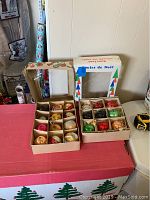 Two open boxes with 24 total vintage glass Christmas ornaments in individual cardboard slots, placed on red and white box with Christmas trees printed.