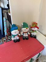 Three standing snowman decorations displayed side by side on a red surface against a white wall, showing their full body and different hats and scarves.