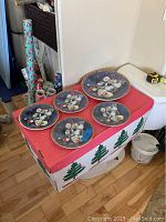 View of all five plates on a red and green decorated box, one large serving plate center with four smaller side plates surrounding it, all decorated with snowman images.
