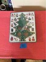 Full box set of wooden Christmas tree and ornaments in original tray packaging atop red surface with rolled wrapping paper in background.