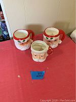 Three ceramic Santa Claus head mugs on a red cloth. Two large mugs and one smaller mug are visible, all featuring red handles and holly decoration.