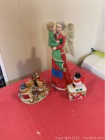 Photo showing all five items: large angel statue holding child, snowman trinket box, and three singing bear figurines arranged on a red cloth.