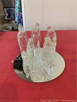 Photo of entire glass nativity set showing seven clear glass figures arranged on round mirror base on red surface.