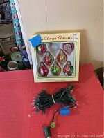 Boxed set of eight hand-decorated glass Christmas ornaments in assorted colors and shapes, on pink table