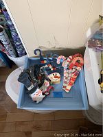 Overview photo of a blue plastic container holding multiple flat, hand-painted wooden Christmas ornaments: snowmen with black top hats and red scarves, candy canes with red and white stripes, and other festive shapes, all with wire hanging loops.