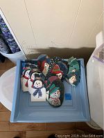 Top-down view of all 21 hand painted wooden Christmas ornaments placed in a storage bin, showing a variety of snowmen and other holiday designs with wire hooks for hanging.