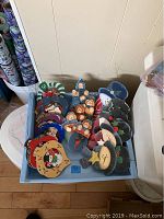 Photo of the lot showing a blue plastic bin filled with multiple hand painted wooden Christmas ornaments including snowmen, bears, and other characters.