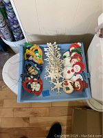 Overview of 25 hand-painted wooden Christmas ornaments including snowmen and snowflakes in a blue plastic bin