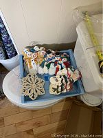 Blue container holding a batch of hand-painted wooden snowmen ornaments and one or two snowflake ornaments visible at the front.