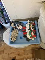 Photo showing assortment of 23 hand painted wooden Christmas ornaments including snowmen, red ice skates, and snowflakes arranged on a blue tray.