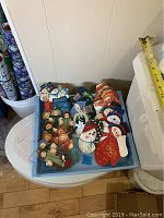 Photo showing assortment of flat, hand painted wooden Christmas ornaments piled in a box, mainly snowmen shapes and designs.
