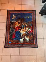 Fabric wall hanging or throw with detailed nativity scene illustrating Mary, Joseph, baby Jesus, shepherds, animals, and star at night, with a colorful border.