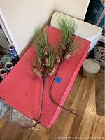 Two artificial tree sprigs with long green pine needles, pine cones, and red berries on a red surface edge, showing full length and detail.
