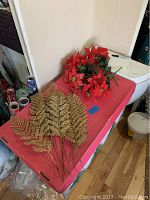 Photo of 10 gold glitter leaf sprigs and 4 bunches of artificial red poinsettia flower sprigs with green leaves and stems on a red surface