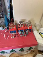 Three ceramic light-up village houses on red surface with boxes behind, plus trees, lampposts, and small ceramic people figurines grouped nearby.