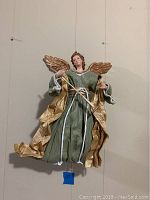 Full view of the angel figurine showing green and gold paper mache robe, gold wings, and white cords.