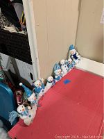 Snowman mantel decor with seven small snowmen figures on white base, arranged on a red cloth surface near a wall.