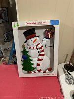 Metal snowman yard stake in box labeled Decorative Metal Pick, showing full front with snowman holding tree and present.