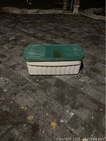 Photo of the large plastic storage bin showing top view with green lid, placed on stone pavement at night.