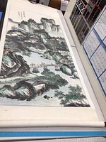 Full view of watercolor scroll painting laid out on table. Shows detailed landscape with mountains, trees, water, bridge, and figures.