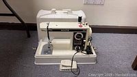 Photo of entire sewing machine with case closed, cord and foot pedal attached, placed on carpeted floor.