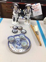 Overview photo of two ceramic Asian figurines, small dishes with dragon design, three small cups, three small decorative pieces, and a hand paper fan on a table.