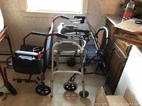 Photo showing one manual wheelchair with black seating and red frame, two walkers including one with knee rest and one stand-up walker, and two walking canes leaning on the wheelchair.