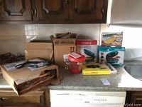 Overview of kitchen counter with boxed items: Holstein waffle maker, brownie maker, Egg & Omelet Wave, cast iron biscuit pan, glass jar, and boxed crepe maker.