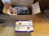 Open cardboard box showing Ninja 4-in-1 cooking system wrapped in plastic inside with instruction manual. Rival food steamer box is positioned outside box on floor.