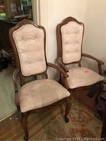 Two wooden dining chairs with carved wooden frames, cane backs, and off white jacquard fabric padded seats and backrests. One has arms, the other does not.
