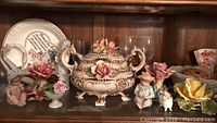 Photo of wood shelf with ceramic soup tureen, glass goblet, figurines and floral decorations
