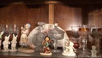 Shelf display of various glassware including clear decanter with glass stopper, multiple etched wine glasses, and porcelain figurines including girl with deer, rabbits, dancing child, and unicorn.
